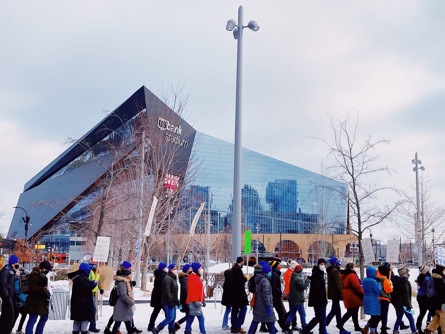 group of protestors walking on the sidewalk carrying signs in front of the us bank stadium