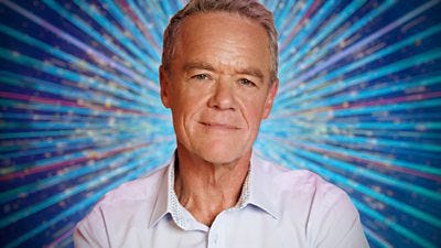 Stefan Dennis smiles to camera in a light shirt, set against a glittering blue background