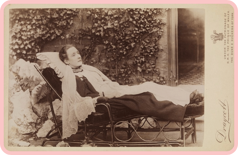 A sepia photo of a woman reclining on a chaise longue, outside of a house. Her neck is propped up with pillows, and she is wrapped up in blankets, her legs outstretched in front of her. She is looking off into the middle distance, with a blank expression on her face. Behind her, an open door leads inside a house, and the exterior walls are covered in ivy. A sepia photo of a woman reclining on a chaise longue, outside of a house. Her neck is propped up with pillows, and she is wrapped up in blankets, her legs outstretched in front of her. She is looking off into the middle distance, with a blank expression on her face. Behind her, an open door leads inside a house, and the exterior walls are covered in ivy.
