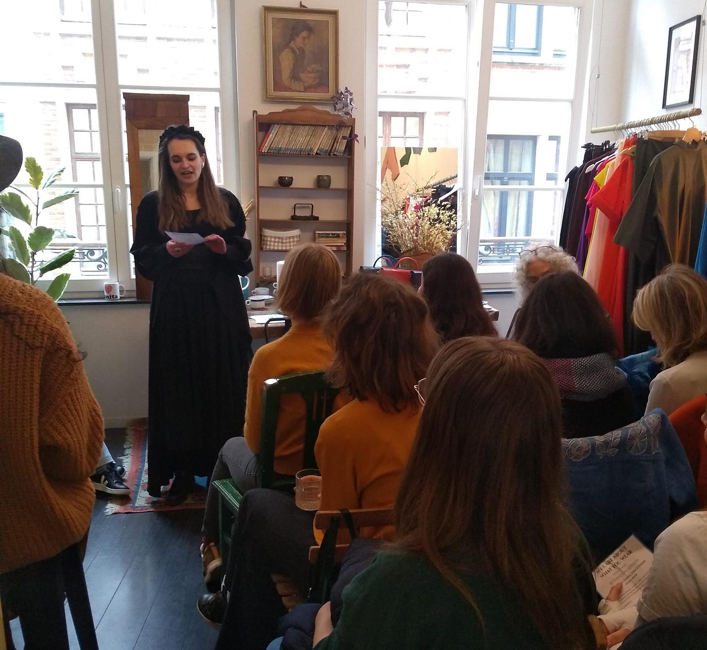 Woman in black dress and black headband speaks to sitting audience of around 15 people in a fashion shop