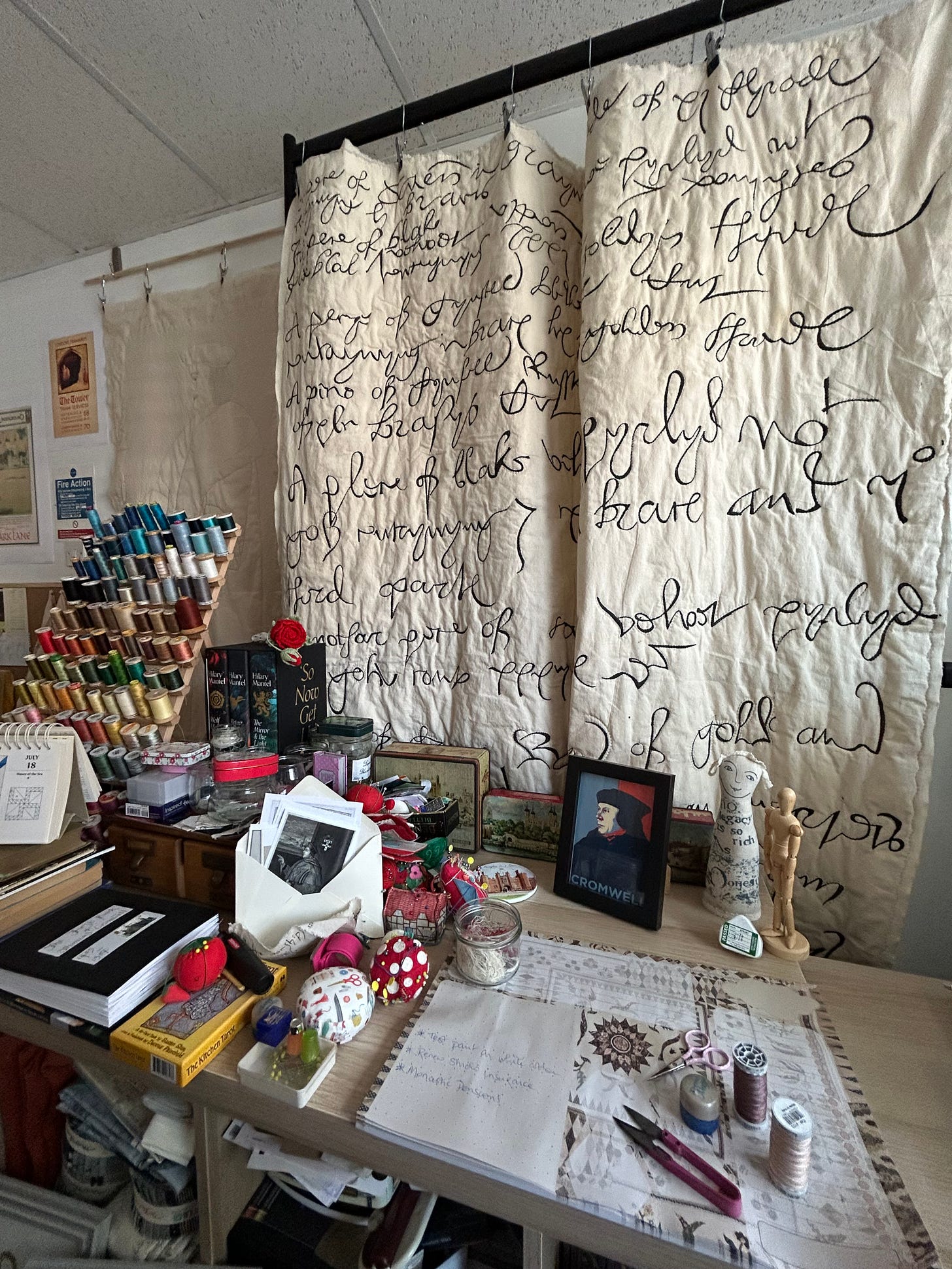 A working space - sewing threads and notions. A portrait - in modernist style - of Thomas Cromwell, and old tins featuring the Tower of London. A large quilted piece of C16th lettering hangs behind the work area. 