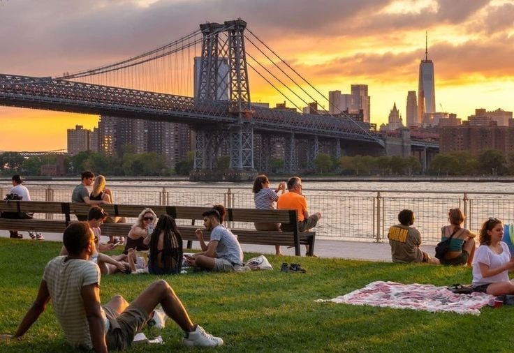 This may contain: many people are sitting on the grass by the water at sunset in front of a bridge