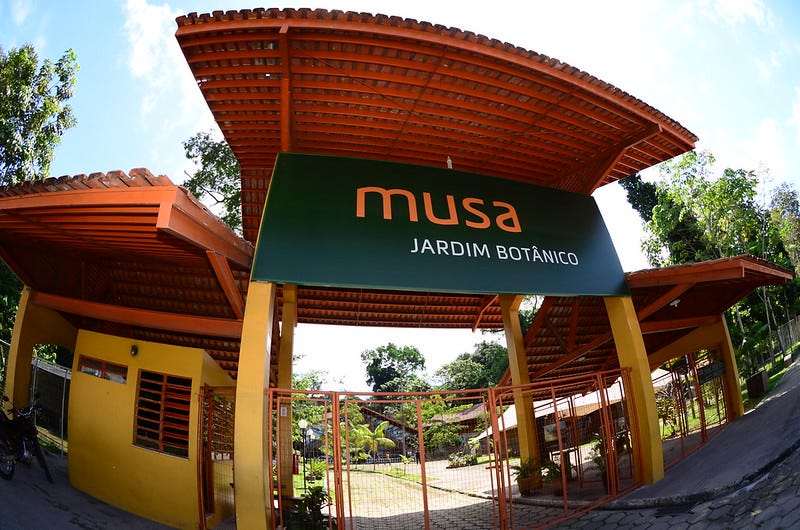 Entrada do Museu da Amazônia. Portão de grade metálica laranja ancorado em pilastras amarelas de concreto, que sustentam um telhado de caibros e terças de madeira e telhas de barro. Placa verde com o nome "musa" em letras laranjas e "jardim botânico" em letras brancas. Do lado de dentro da área do museu é possível observar as árvores.