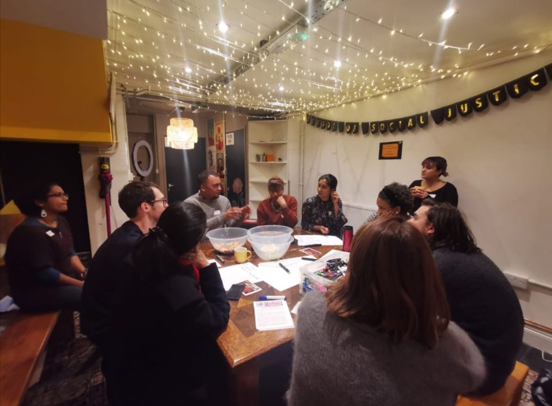 A group of people gather around a table for a meeting with paper, listening to one man speak