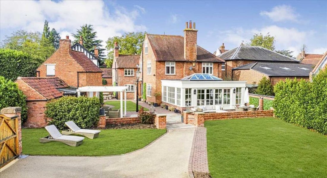 A large brick farmhouse with a white conservatory, canopy, sun loungers, and a lawn in front of it