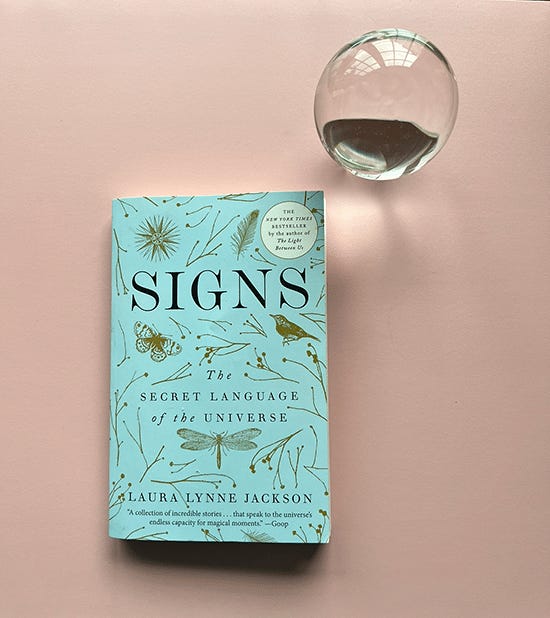 The book “Signs” sits against a blush pink background. A crystal orb sits next to it. The book “Signs” sits against a blush pink background. A crystal orb sits next to it.