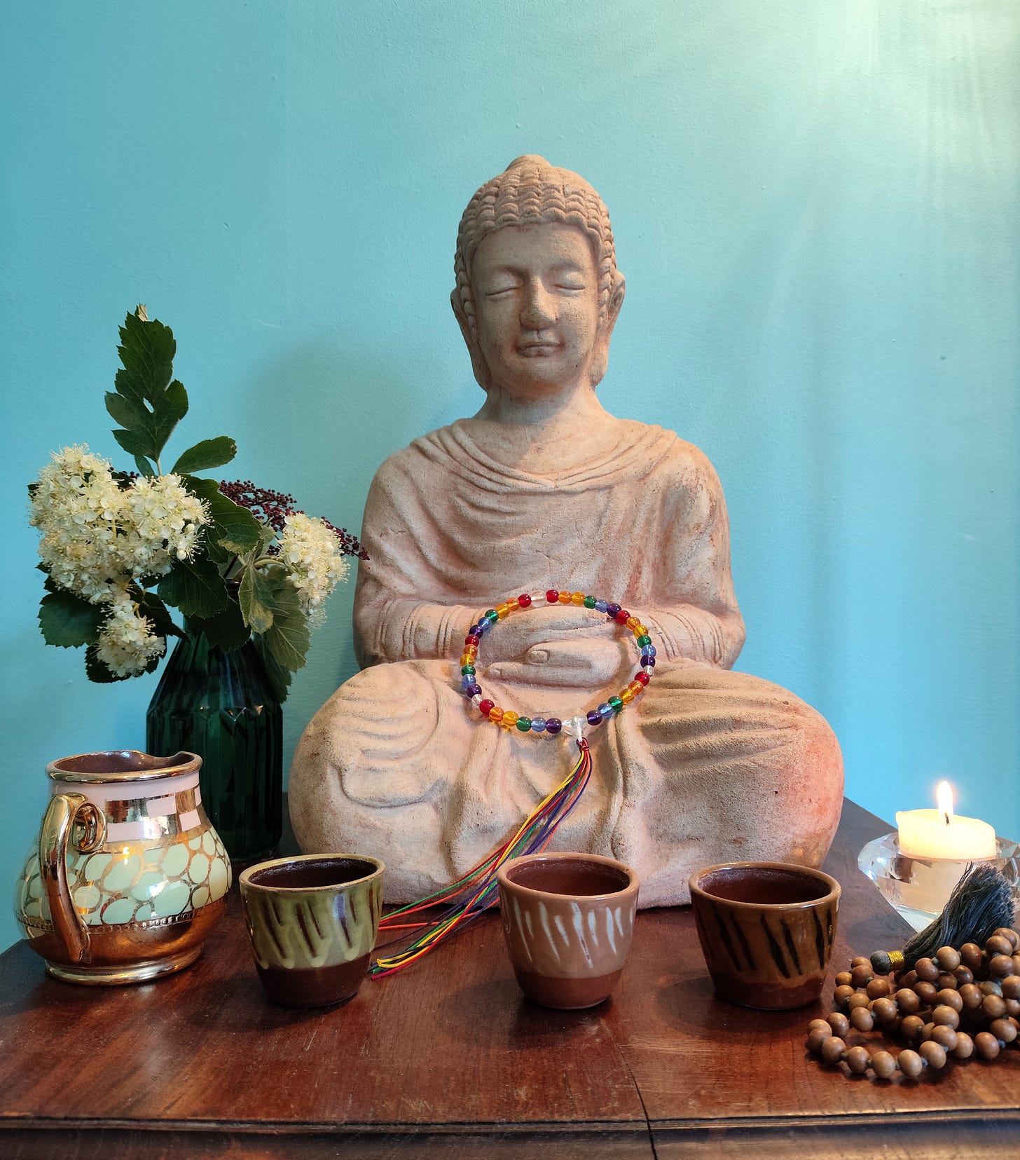 A stone buddha on a wooden table. In front of the Buddha are three clay cups, there is a small jug to the left of the cups, and a mala to the right. There is a candle to the right of the Buddha, and a vase with flowers to the left.