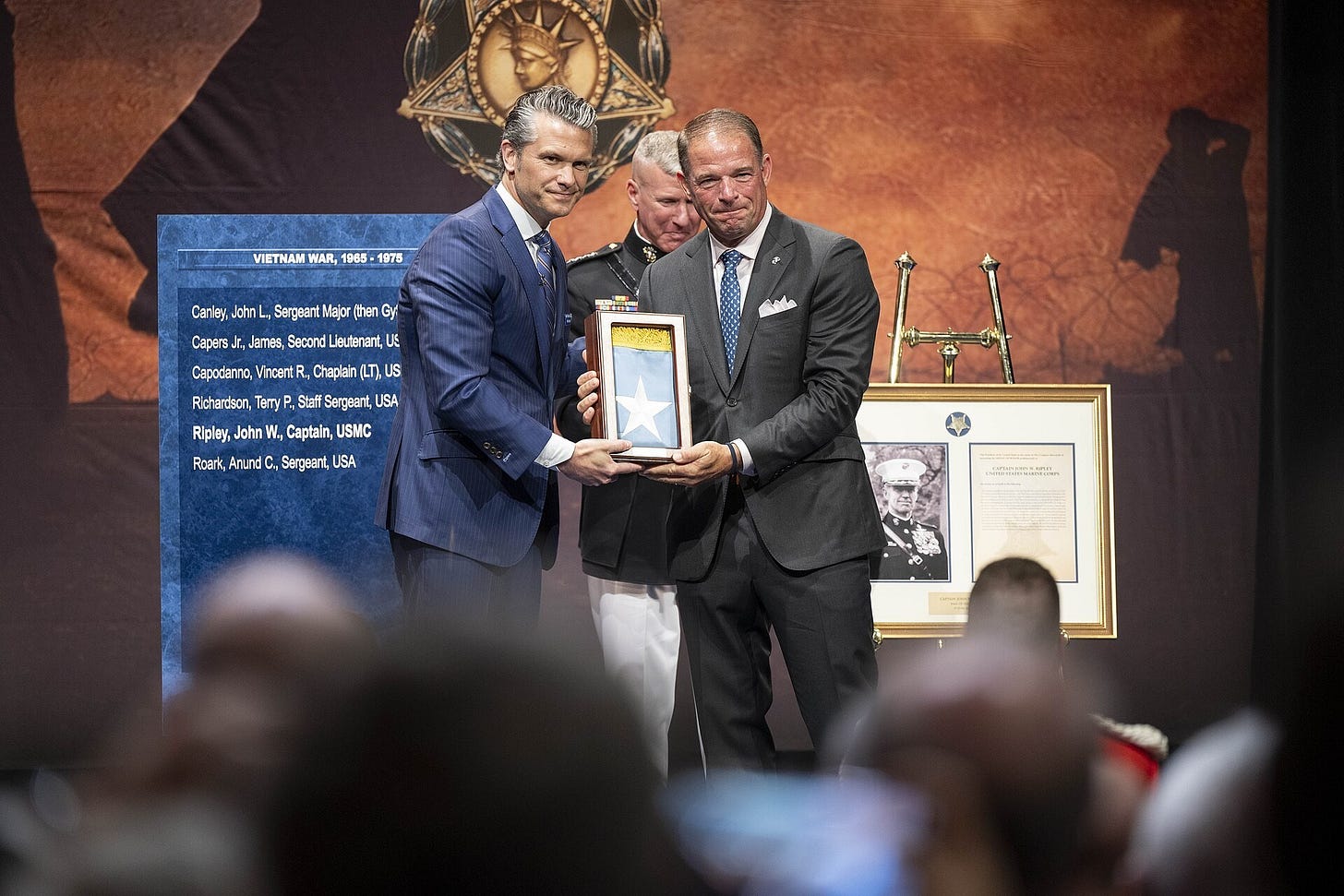 Two men in suits exchange a framed citation on a stage in front of a Medal of Honor backdrop and a blue panel listing names. Two men in suits exchange a framed citation on a stage in front of a Medal of Honor backdrop and a blue panel listing names.