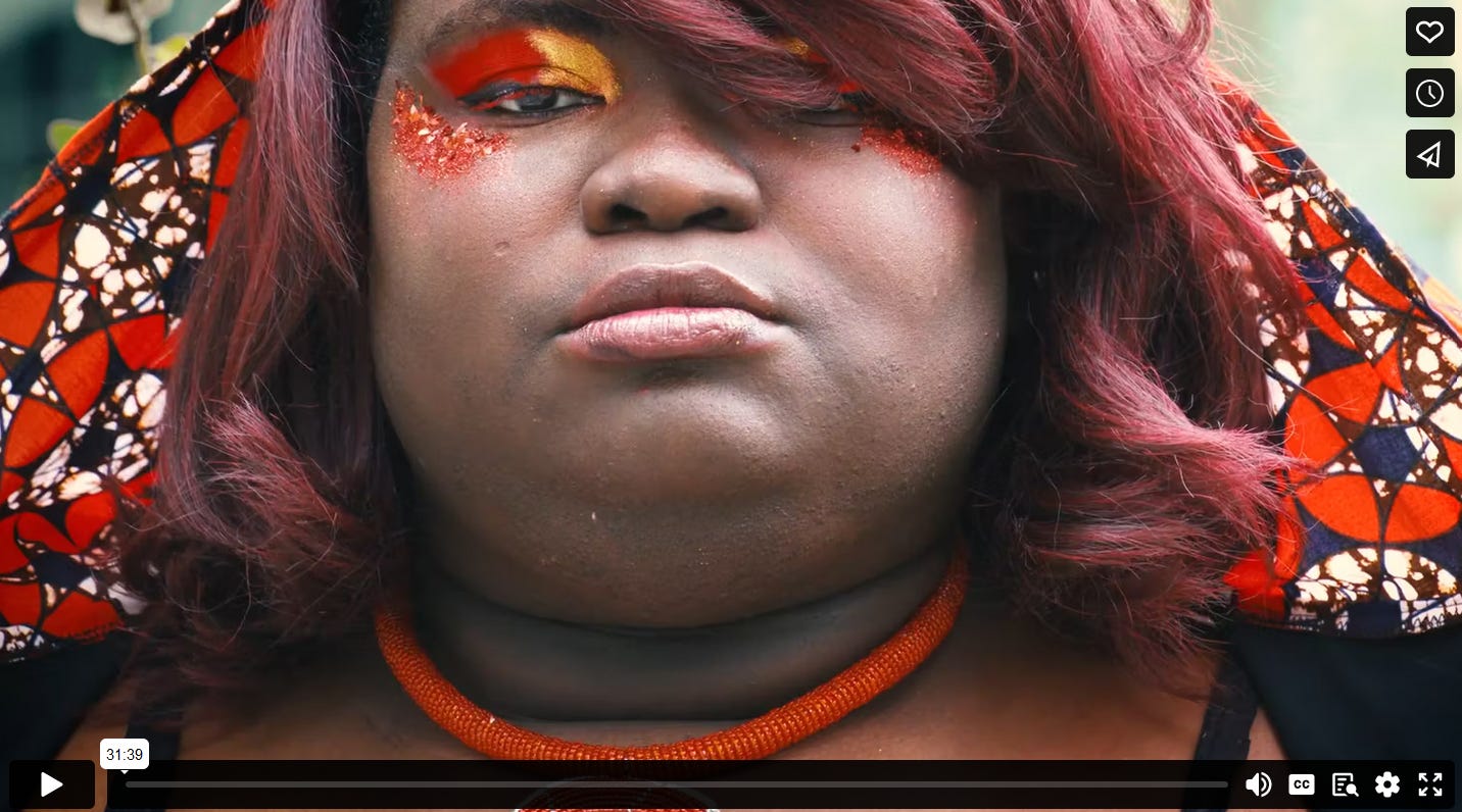 Close-up on the face of a Black model; she is wearing a hooded cape with a red, blue, and white geometric print and a red beaded necklace. She also has bright red and gold eyeshadow and glitter. 