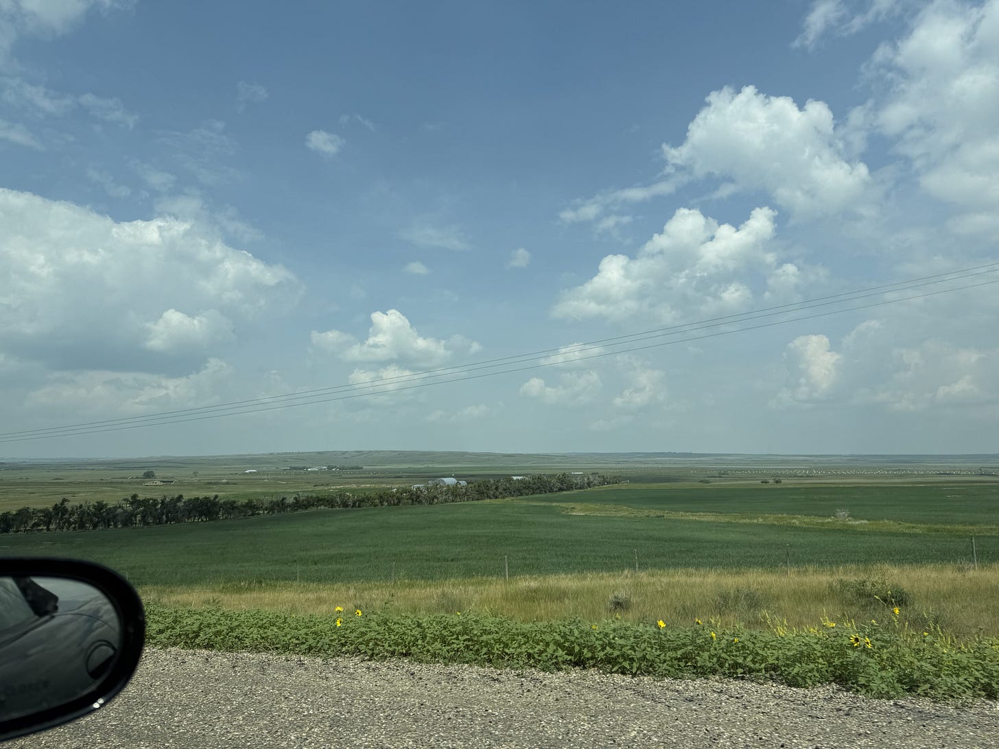 A scenic view from a car featuring a blue sky with fluffy clouds, green fields, and distant hills.