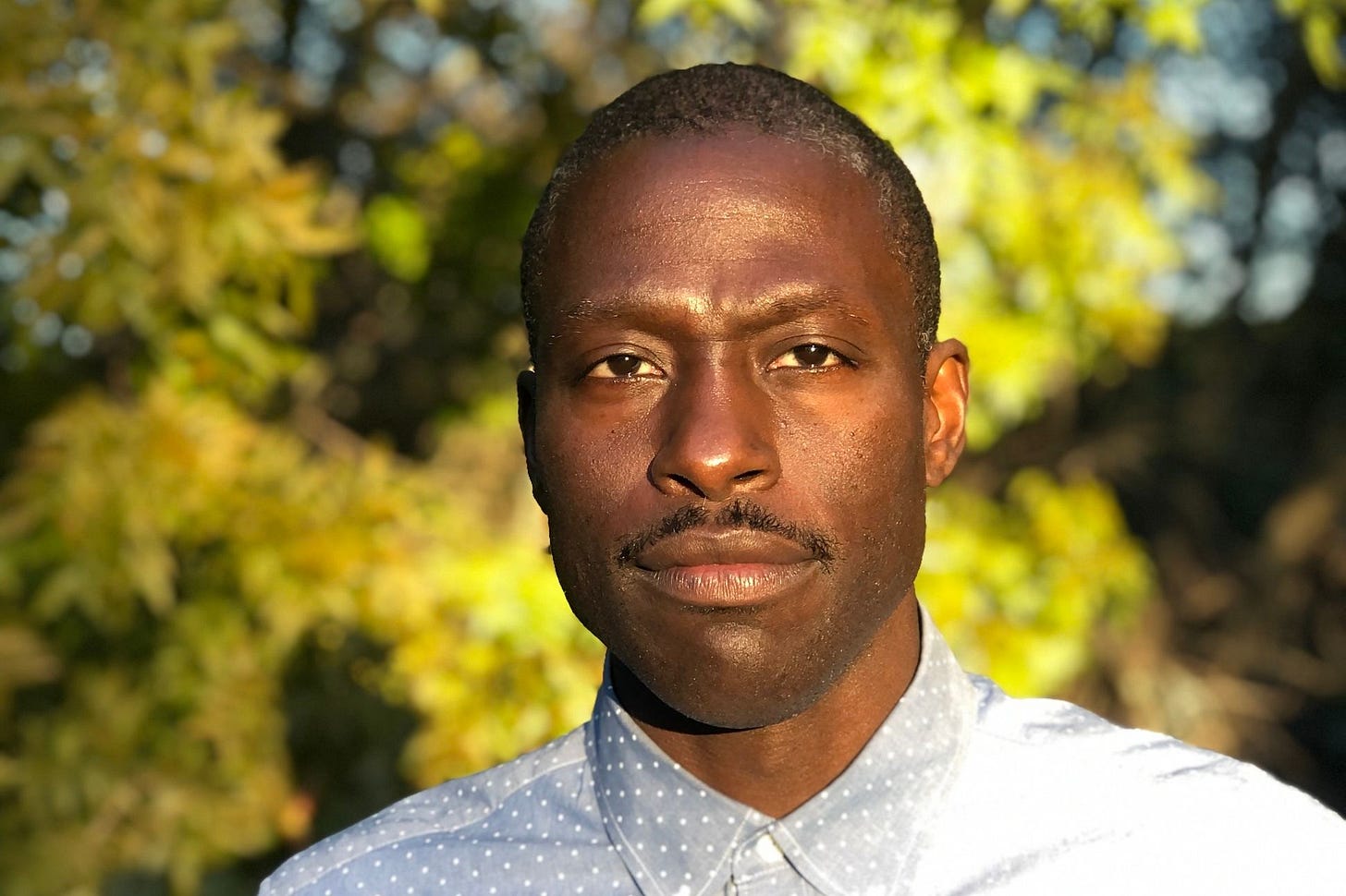 color headshot of Roger Reeves wwearing a light colored shirt. In the background, blurred, are summer or early-fall trees