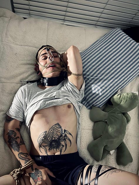 A collage of photos of Rae in various forms of bondage: ranging from chained to a car hoist, to rope and rope suspension as well as cuffs and leather bound to a bed, or their ankles. Rae has many tattoos on their arms, legs and stomach. They have a shaved head and brown eyes. They have a large septum ring, a bridge piercing, and stretched earlobes.