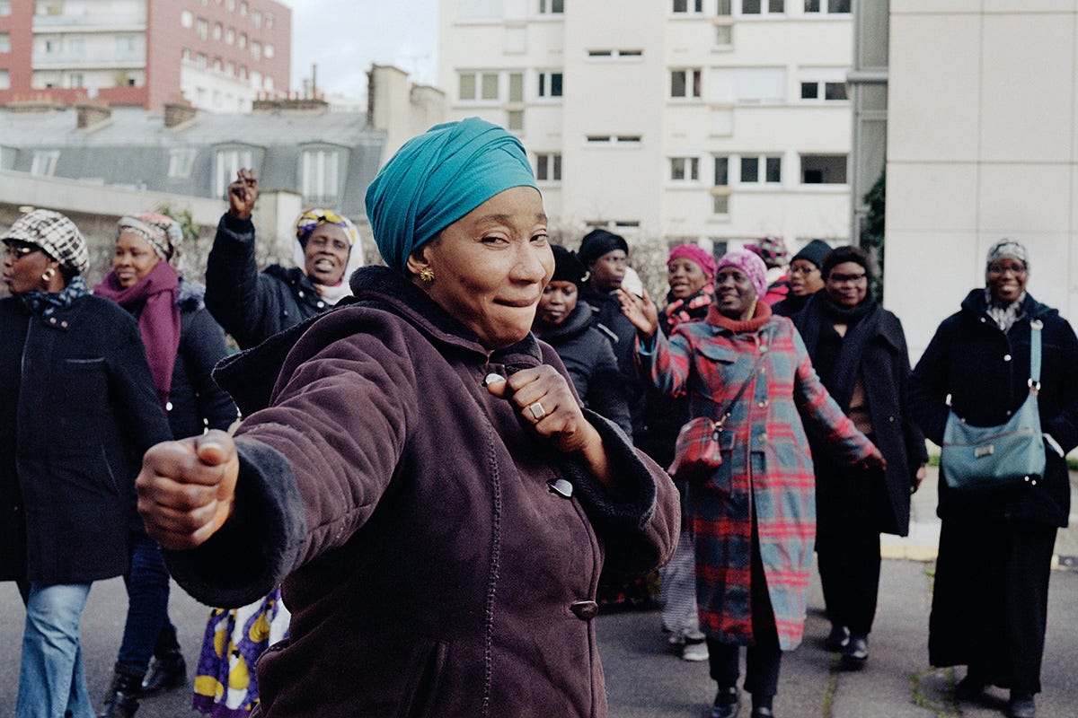 A Black African woman wearing a headwrap, surrounded by other Black African women in the streets of Paris, winks at the camera as they appear to be part of an organised labour protest. A Black African woman wearing a headwrap, surrounded by other Black African women in the streets of Paris, winks at the camera as they appear to be part of an organised labour protest.