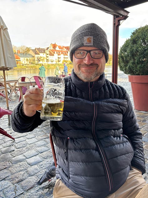 a gothic cathedral, man holding a mug of beer, a plate of sausages and bread, building by a river, a wall of cuckoo clocks, closeup of a christmas musical piece in the cuckoo shop