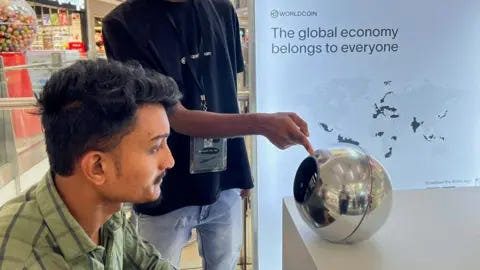 Reuters A man in India sitting before an earlier World device, a silver orb, to have his eye scanned as a form of human identification. He has dark hair and a green shirt, and looks straight into the orb, on a desk in front of him, while another man points to it. 