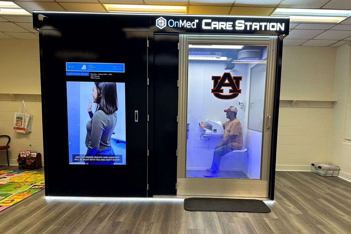 A booth with a large screen on the left and a door with a window on the right and the text "OnMed Care Station" at the top. Through the window can be seen a man in a t-shirt, jeans, and cap sitting inside a booth with his right arm in a device. A booth with a large screen on the left and a door with a window on the right and the text "OnMed Care Station" at the top. Through the window can be seen a man in a t-shirt, jeans, and cap sitting inside a booth with his right arm in a device.
