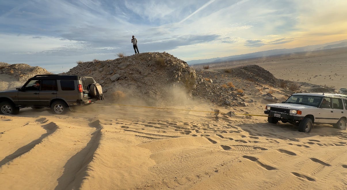 A white Land Rover Discovery pulls another Land Rover up a sandy desert hill during an off-road recovery. Dust kicks up around the vehicles while a person stands on a rocky ridge above watching the recovery unfold against a wide desert landscape and cloudy evening sky. A white Land Rover Discovery pulls another Land Rover up a sandy desert hill during an off-road recovery. Dust kicks up around the vehicles while a person stands on a rocky ridge above watching the recovery unfold against a wide desert landscape and cloudy evening sky.
