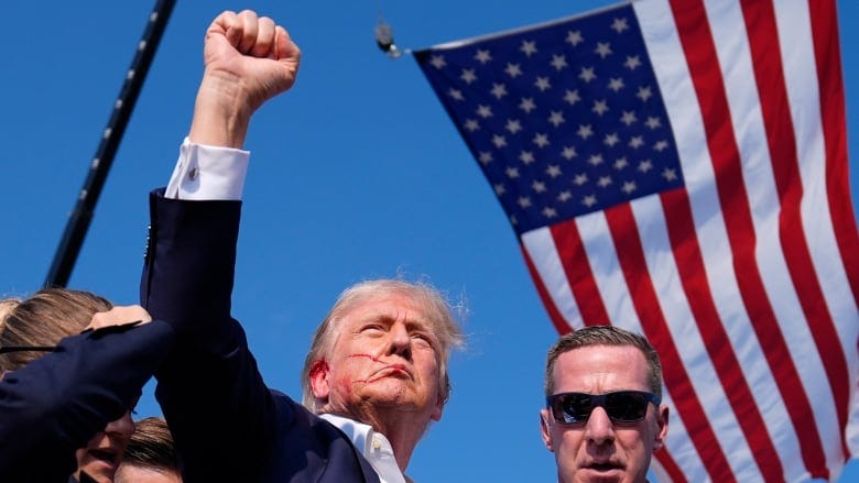 A clean-shaven older man with blood on his face raises his right fist while surrounded by a handful of people who, like him, are wearing dark suit jackets.
