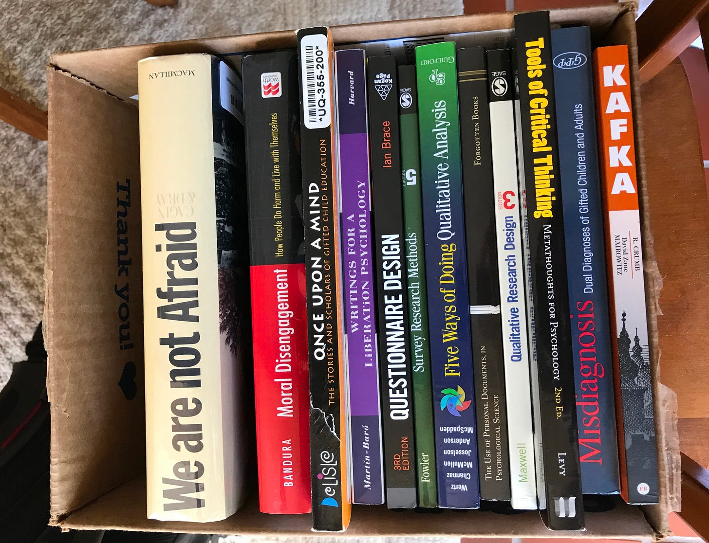A cardboard box filled with books brought from Colorado for a 2017 research trip. The visible titles include We Are Not Afraid, Moral Disengagement by Albert Bandura, Once Upon a Mind, Writings for a Liberation Psychology, Five Ways of Doing Qualitative Analysis, Narrative Research Design, Tools of Critical Thinking, Misdiagnosis and Dual Diagnoses of Gifted Children and Adults, and Kafka.