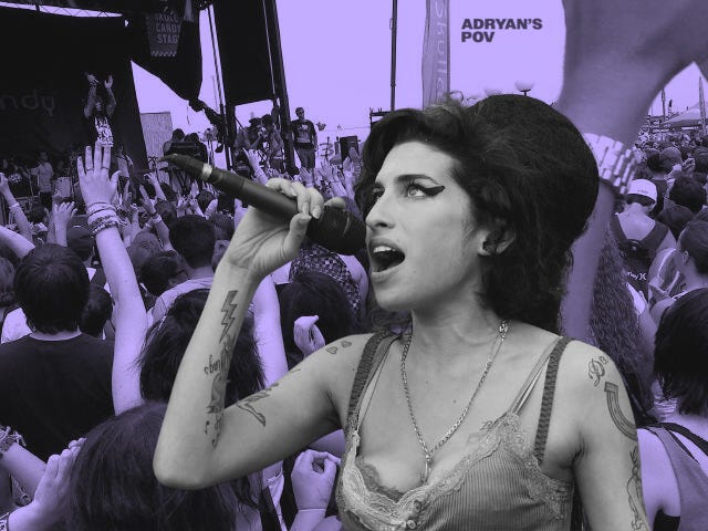 A digitally altered photo of Amy Winehouse singing into a microphone at an outdoor music festival. She is wearing a tank top with lace trim, a long necklace, and has her signature beehive hairstyle and winged eyeliner. Her tattoos are visible on her arm, and she appears to be passionately performing. The background shows a crowd raising their hands toward the stage, with some wearing wristbands and festival attire. The image has a desaturated, purple-tinted color overlay.