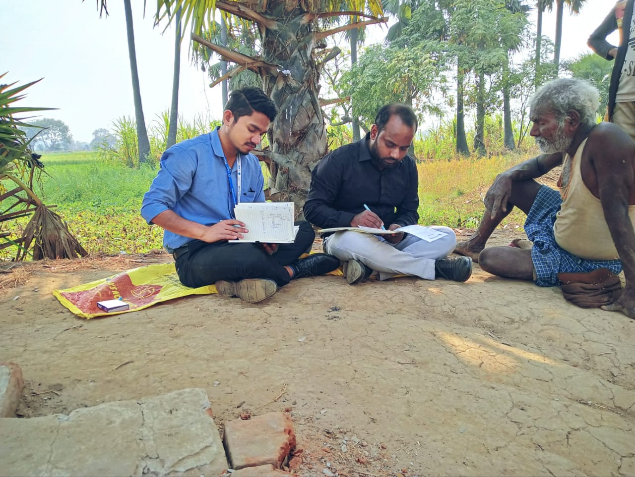 Three people sitting on the ground with the backdrop of a field. Two of them are team law foundation and are interviewing an old farmer Three people sitting on the ground with the backdrop of a field. Two of them are team law foundation and are interviewing an old farmer