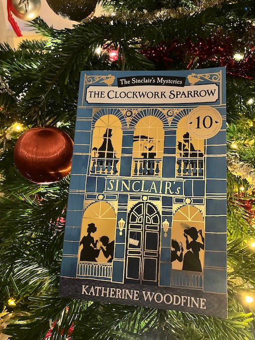 A photograph of the 10th anniversary edition of The Clockwork Sparrow, with a blue and gold cover, and a golden 10th anniversary roundel. The book is in front of a Christmas tree decorated with fairy lights and red and gold decorations.