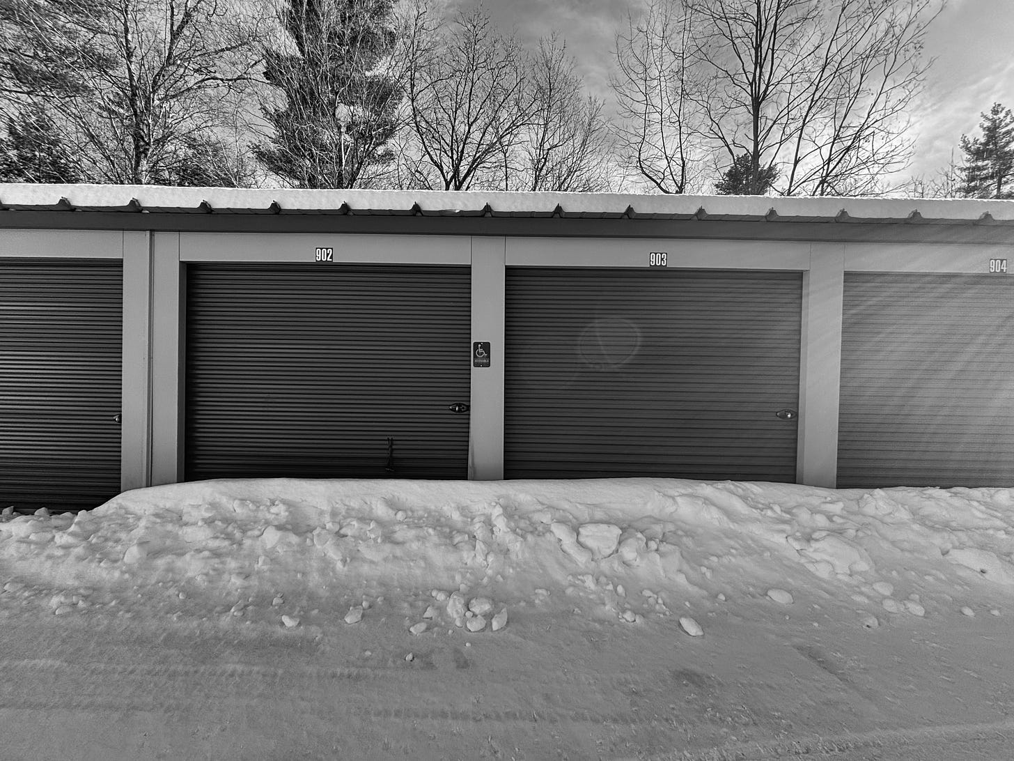 A black and white photo of a row of outdoor storage units surrounded by snow. Trees and sky in the background. A small snowbank built up around the doors to the storage units with a sign above that shows the universal symbol for wheelchair and the word "Accessible."