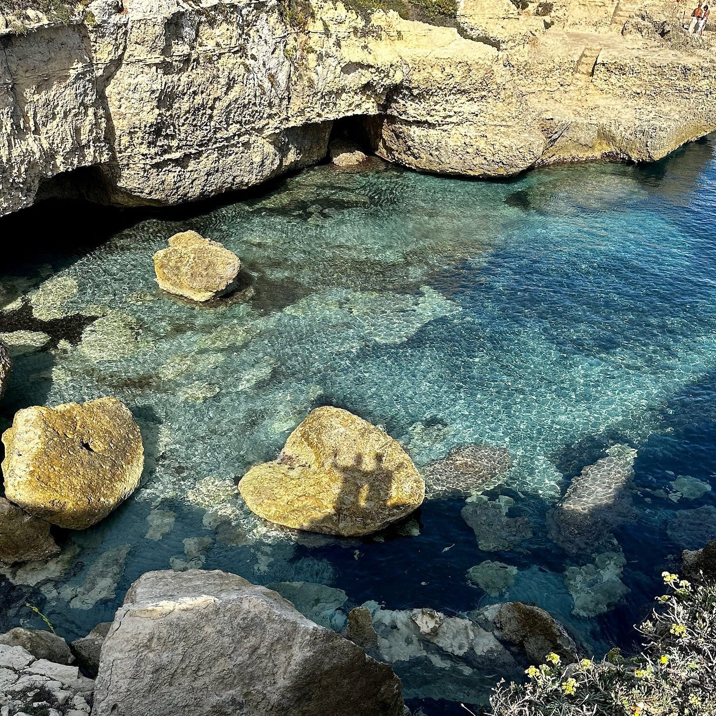The shadow of Kelly and Nigel Benthall cast over turquoise rock pools in Puglia, Italy, reflecting their journey and connection.