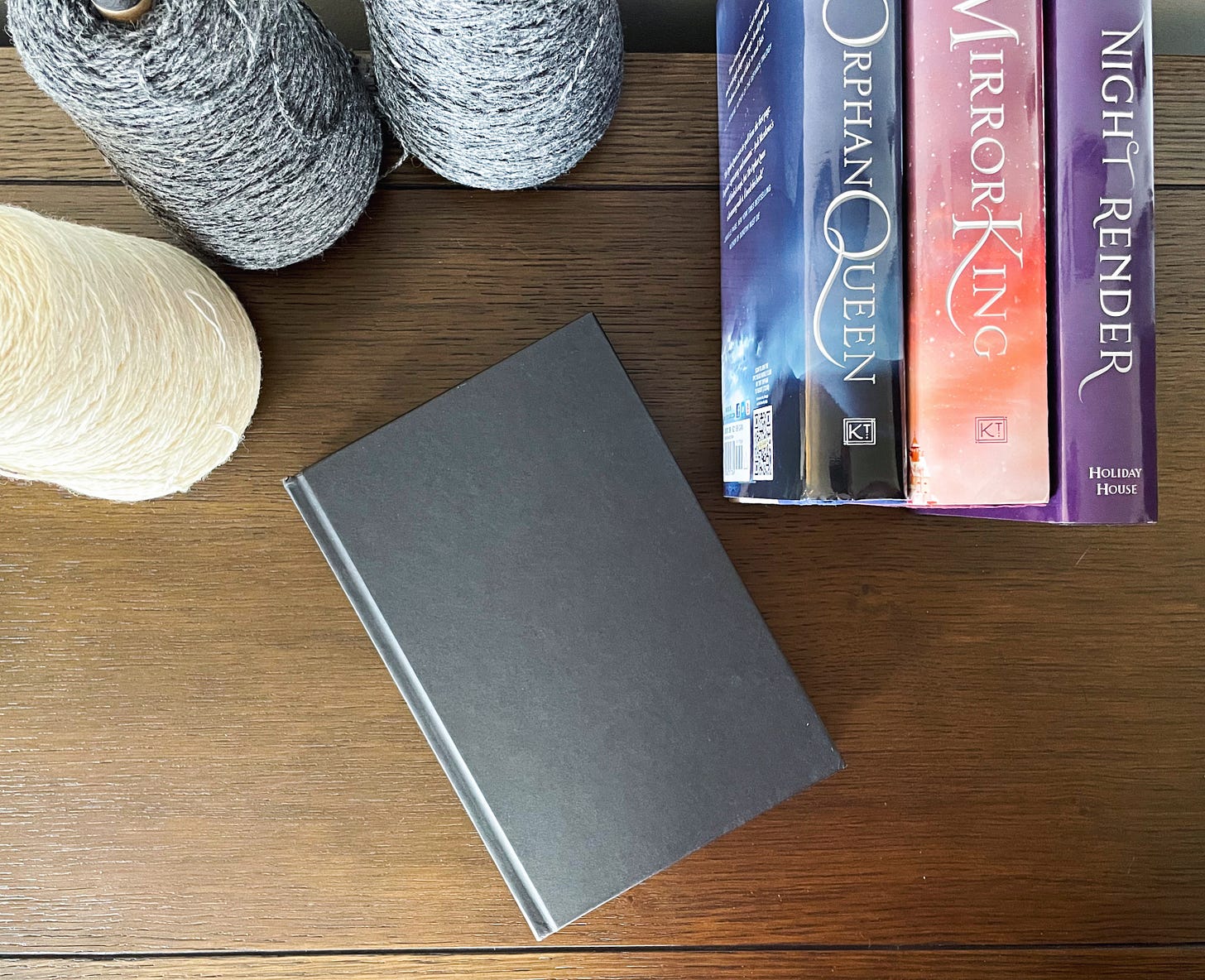 Three spine up books (Orphan Queen duo and Nightrender) and one flat book with no jacket on. Brown background. Three cones of yarn - two gray and one white - on the upper left side.
