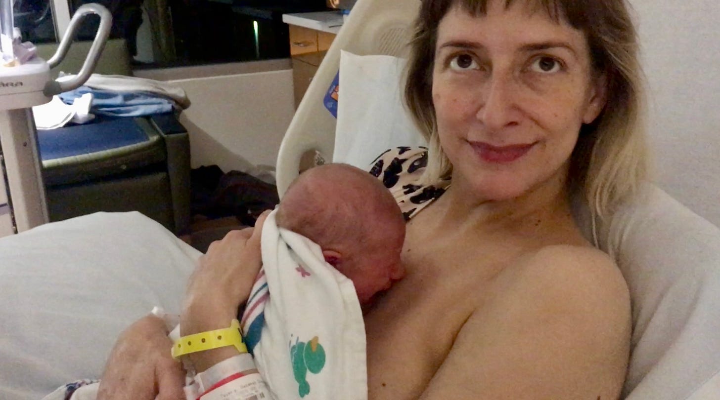 A screenshot from the video described above. Rebekah’s tiny son is crying against her chest, his face red, while she tries to sooth him by patting him on the back. She’s looking up toward her partner who’s holding the camera. Her mouth is smiling and her eyes are pleading. 