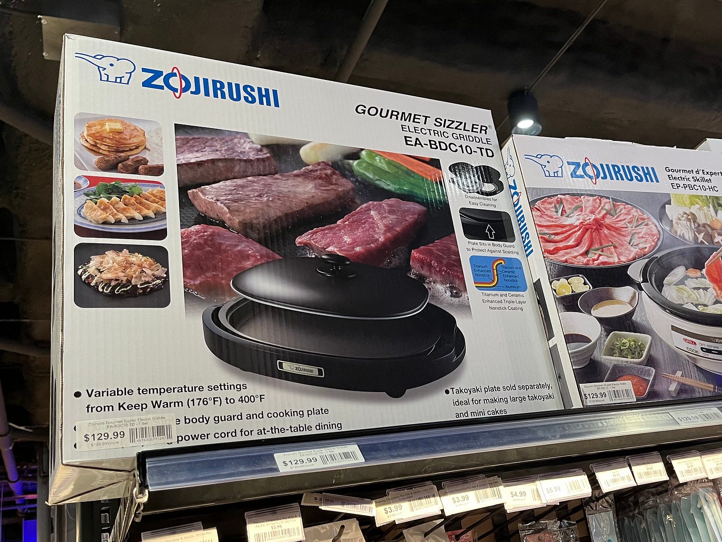 A box of Zojirushi Gourmet Sizzler electric griddle, displayed on a shop shelf. The box shows a photo of the griddle with meat and vegetables being cooked on it. A box of Zojirushi Gourmet Sizzler electric griddle, displayed on a shop shelf. The box shows a photo of the griddle with meat and vegetables being cooked on it.
