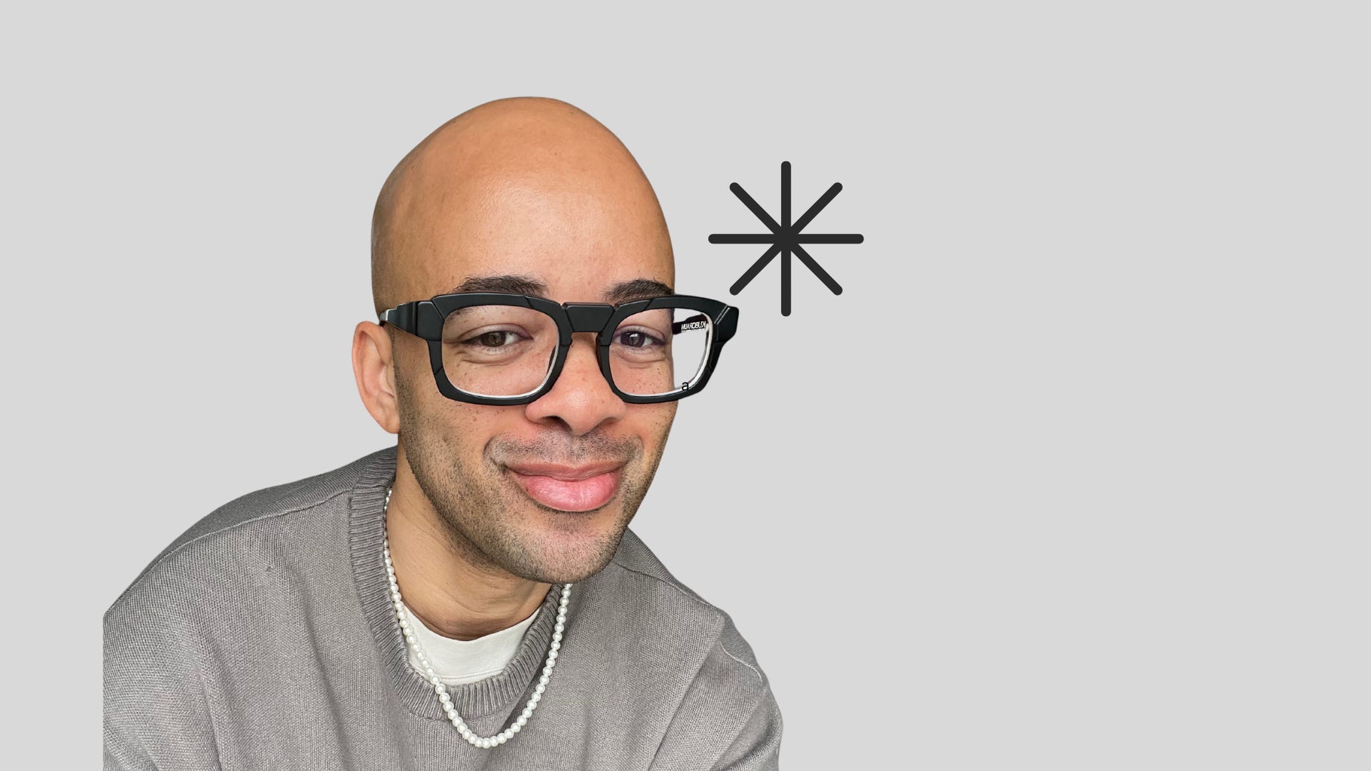 Portrait of Ethan wearing glasses, a sweater, and a pearl necklace, smiling slightly against a light gray background with a black asterisk near his head. Portrait of Ethan wearing glasses, a sweater, and a pearl necklace, smiling slightly against a light gray background with a black asterisk near his head.