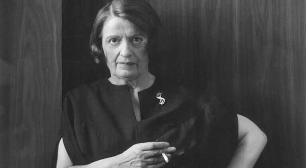 Ayn Rand posing for a portrait holding a cigarette, wearing a black dress and a pin with a dollar symbol.