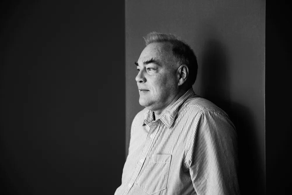 A 2015 black and white photograph of Warren Robinett. He has big bushy eyebrows and he wears a striped button down shirt.
