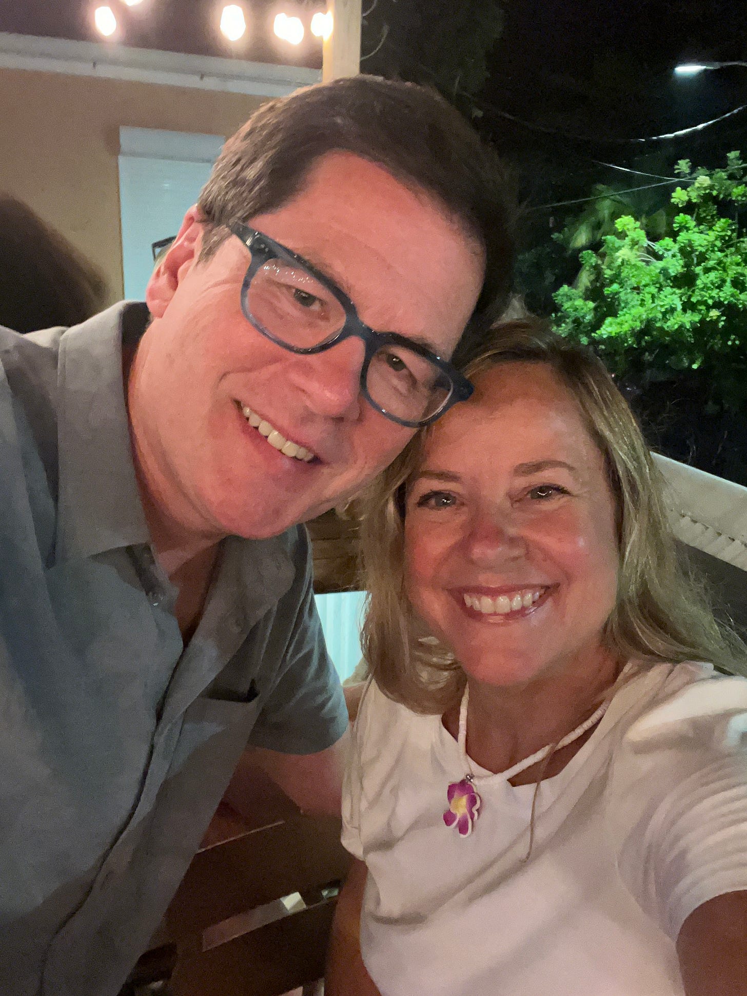 Couple leans heads together for a photo. Man has brown hair, glasses, and a gray shirt. Blonde woman has white shirt and a floral necklace.