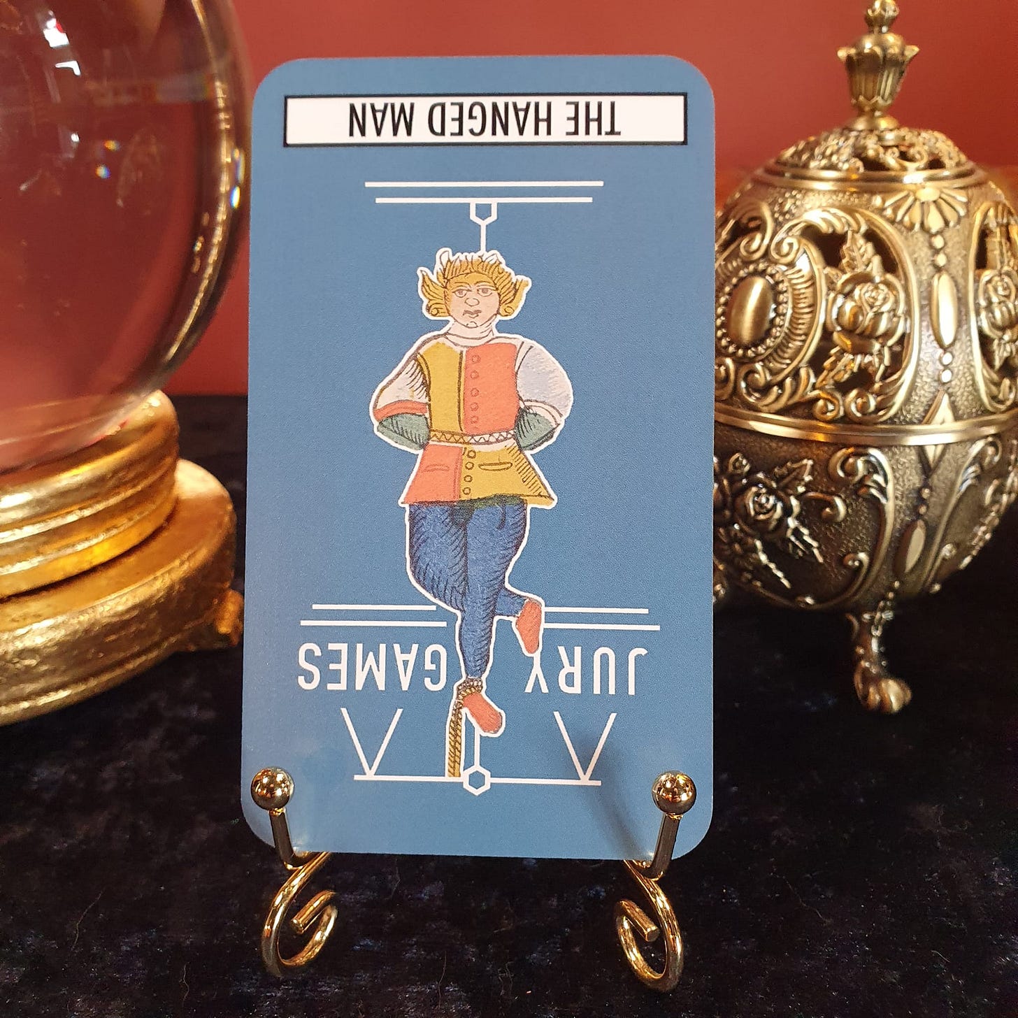 Reversed Hanged Man tarot card displayed on a brass stand beside a crystal ball and decorative incense holder, showing a figure hanging upside down from a beam - photographed for the Tarot DMs interview with Joanne Harris exploring sacrifice, perspective, and tarot symbolism.