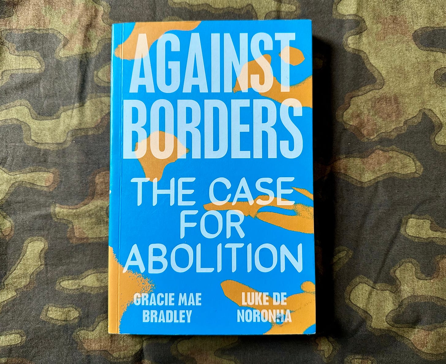Image of the book "Against Borders" by Gracie Mae Bradley and Luke De Noronha on a dark green camouflage fabric. The cover of the book is bright blue with the title taking up most of the cover in large capital letters. Image of the book "Against Borders" by Gracie Mae Bradley and Luke De Noronha on a dark green camouflage fabric. The cover of the book is bright blue with the title taking up most of the cover in large capital letters.
