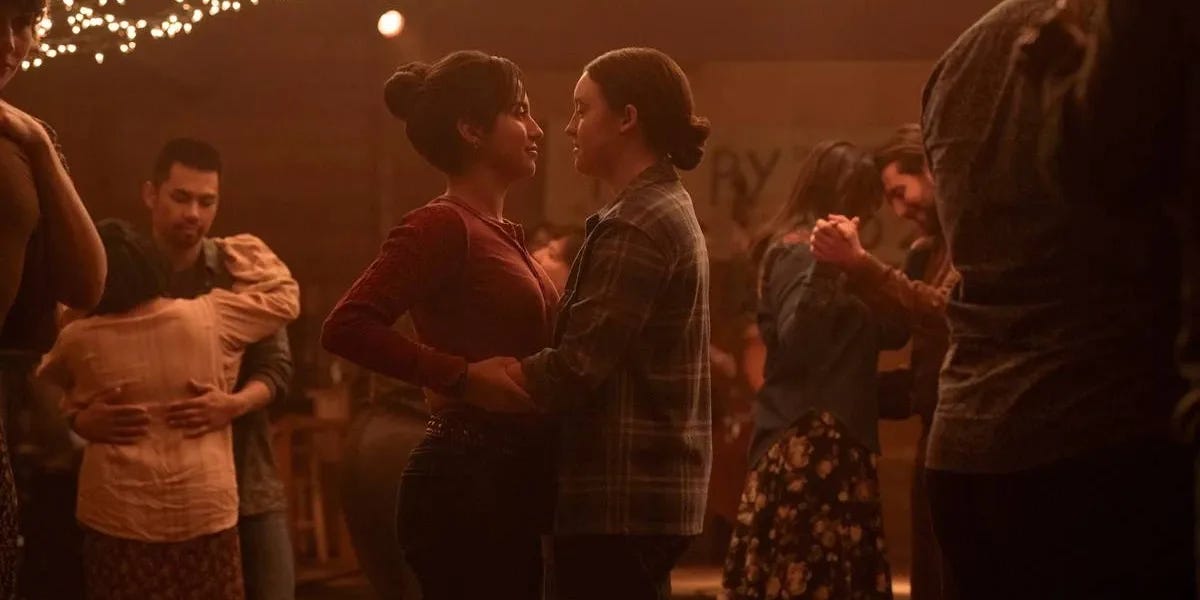 Cena do primeiro episódio da segunda temporada de The Last of Us: Em uma festa, pessoas dançam em pares sob luz baixa, ao centro duas moças dançam juntas, as personagens Dina e Ellie, interpretadas por Isabela Merced e Bella Ramsey, respectivamente