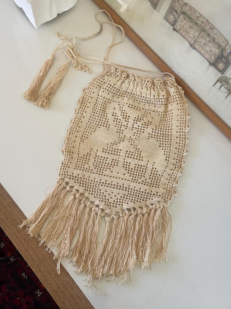 May include: A beige crochet drawstring pouch featuring an intricate openwork pattern with a central motif. The bag includes a long cord with two decorative tassels at the ends and a row of long, dangling fringe along the bottom edge.