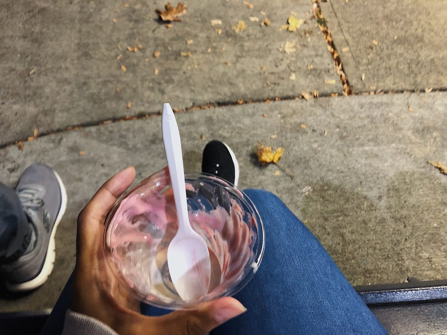 An overhead view of an empty cup of ice cream, and a hand holding it. Beneath the hand and empty plastic cup is a pair of jeans and shoes against concrete