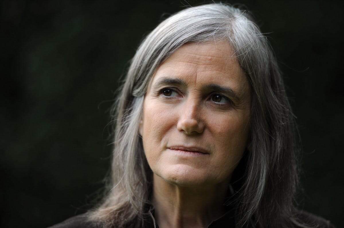 A middle-aged woman with straight gray hair, wearing a dark top, looks thoughtfully into the distance outdoors. A middle-aged woman with straight gray hair, wearing a dark top, looks thoughtfully into the distance outdoors.