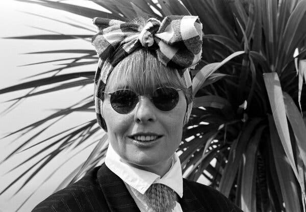 Diane Keaton in round sunglasses standing in front of a plant.