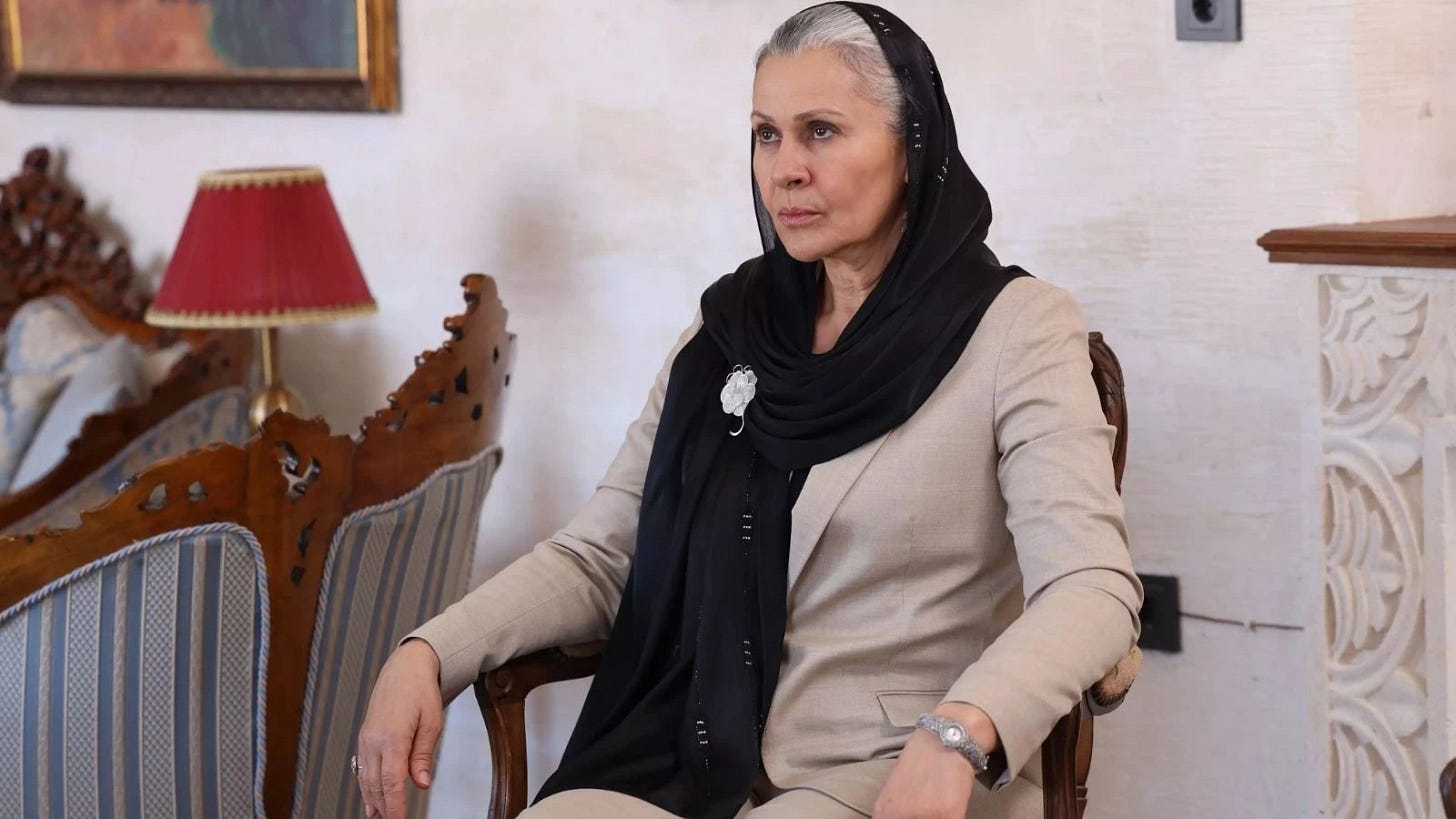 Gonca Cilasun'un siyah başörtüsü ve bej rengi ceket giymiş halde, geleneksel ve zarif bir mekânda oturduğu karakter fotoğrafı. Ahşap oymalı mobilyalar ve mavi desenli koltuk döşemesi önünde, bileğinde gümüş bir saat ile resmedilmiş.