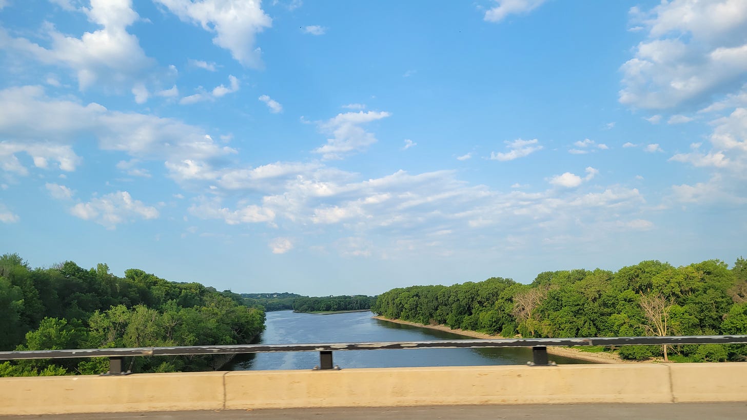 A blue sky with lots of white clouds over the Mississippi River with lush greenery on both banks as seen from a bridge through a car window.