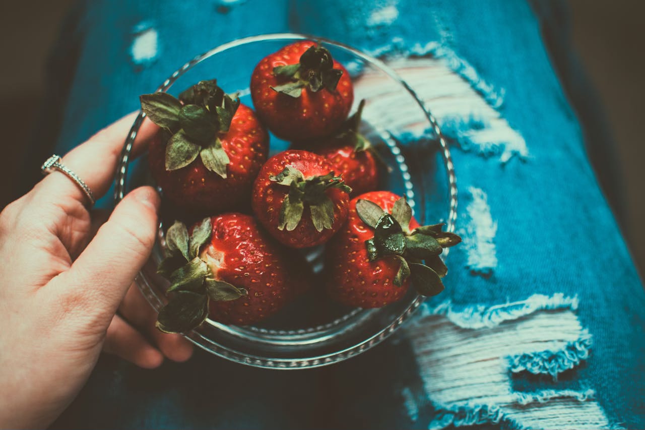A cropped image of a hand holding a clear blow of strawberries on top of a woman's jean-clad legs.