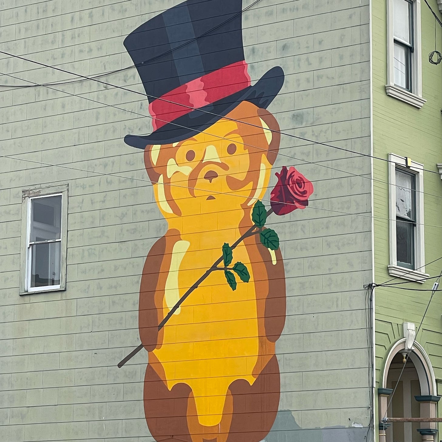 A large mural of a golden honey bear painted on the side of a green building. The bear wears a tall black top hat with a red band and holds a single red rose across its chest.