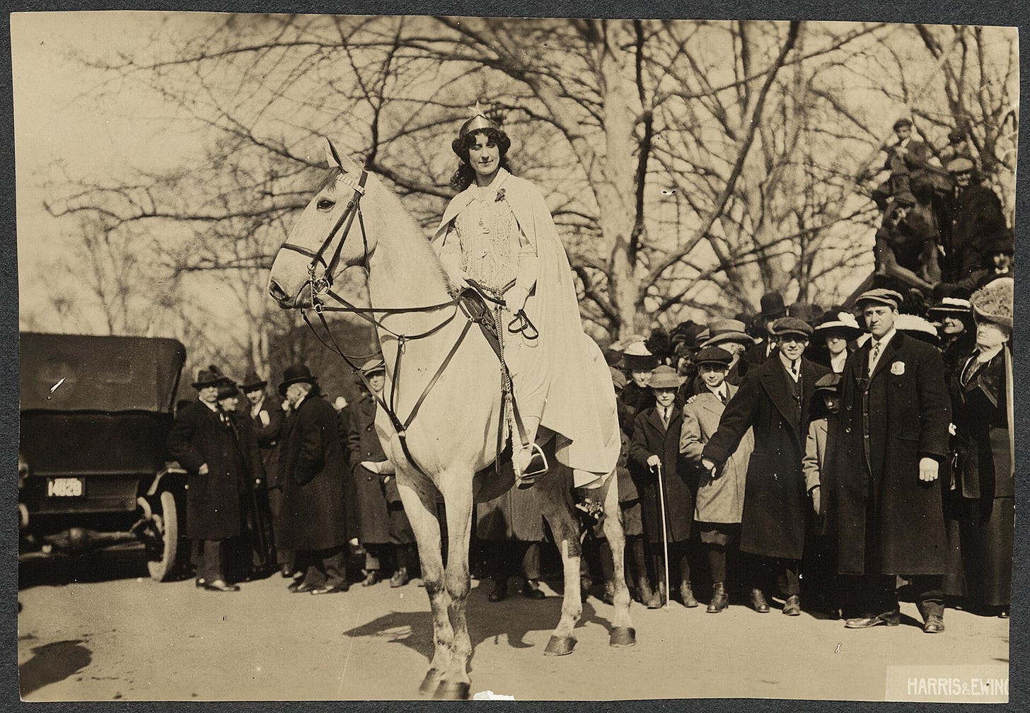 Inez Milholland Boissevain preparing to lead the March 3, 1913, suffrage parade in Washington, D.C. She is wearing white robes and a crown astride a white horse in front of a crowd on a street in Washington, D.C.