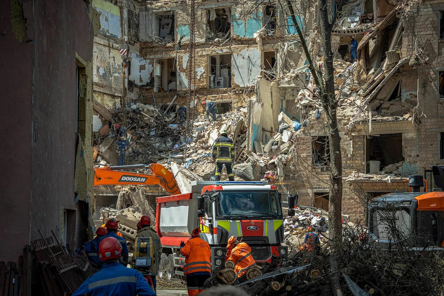 Rescue workers donning orange attire work next a rescue truck outside of a multilevel building that has been hit by a Russian missile and is now crumbling. Debris from the building's wall and the rooms inside has spilled out into the courtyard where the truck is parked.