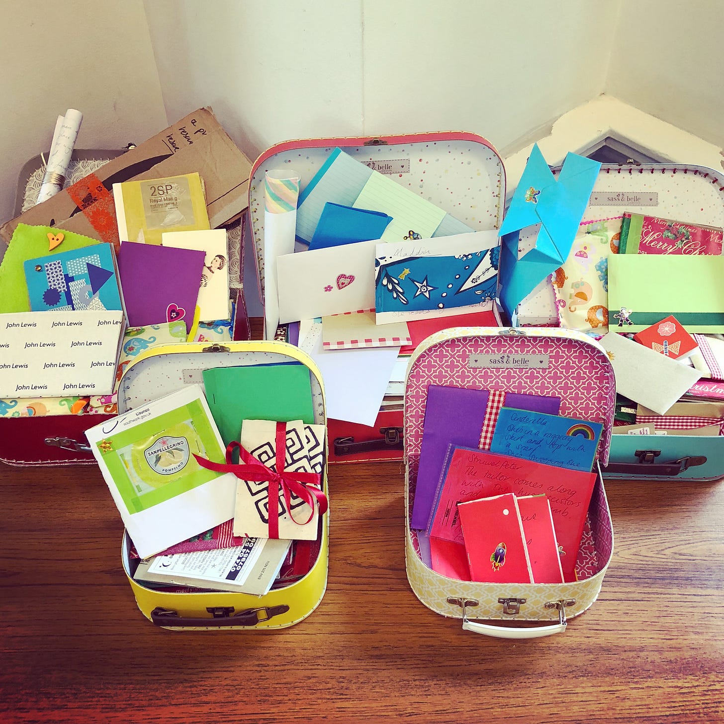 Photo of five tiny cardboard suitcases, each one a different bright colour, stand open and display a collection of scrappy handmade books, some with stickers on, some tied with ribbon, all higgledy-piggledy.