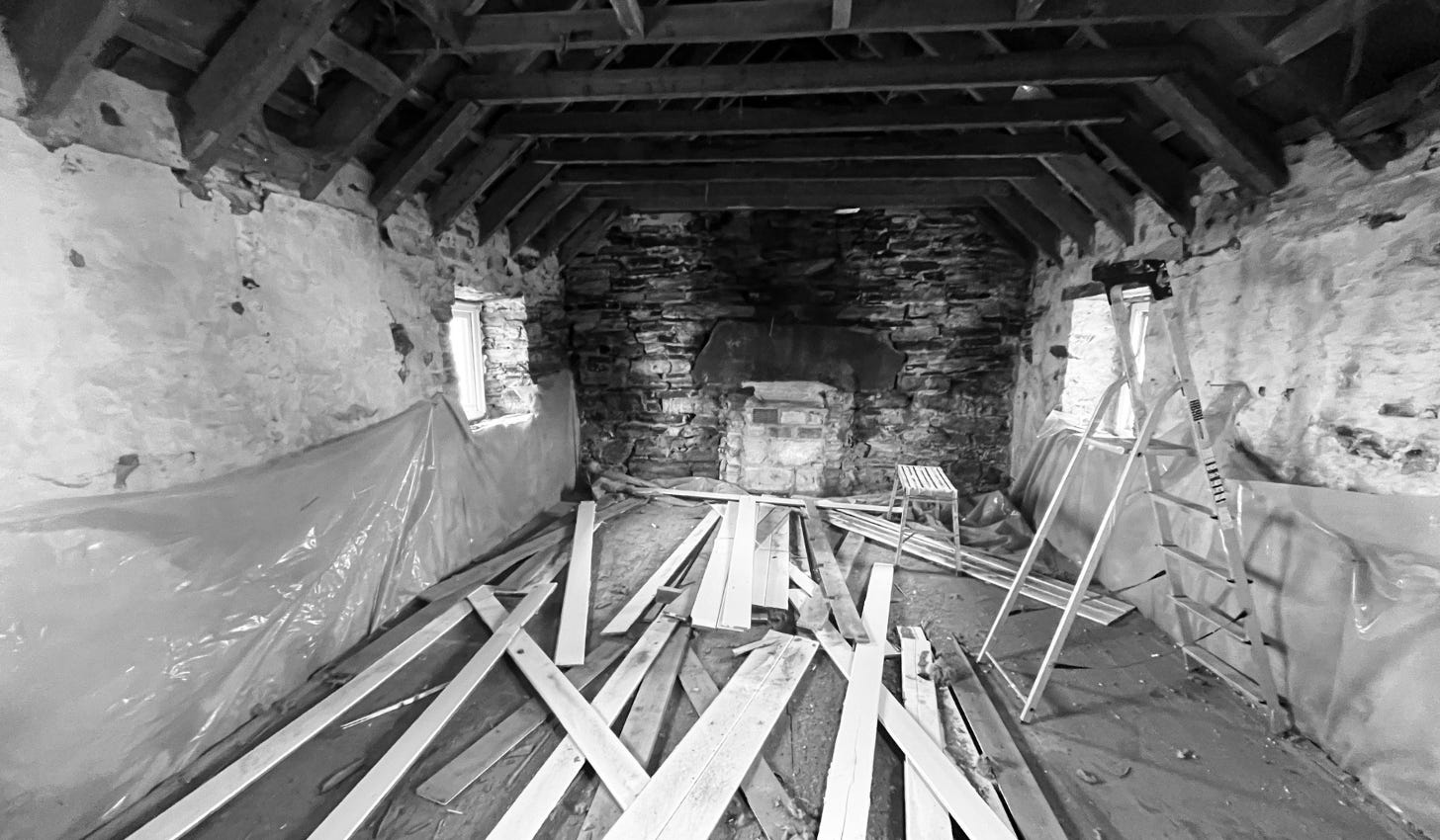 Image of a very sad looking room in B&W - the walls are bare stone & the roof beams are now exposed as the ceiling panels lay in a mess on the floor